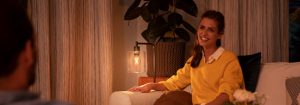 "Acogedor ambiente de salón creado por una cálida lámpara LED de sobremesa, que ilumina a una mujer con jersey amarillo disfrutando de una conversación vespertina".