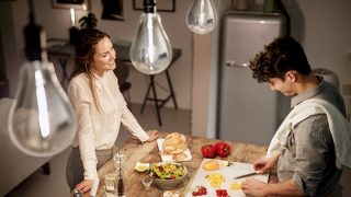 Una mujer observa a un hombre cortando vegetales en una tabla de cortar en una cocina; se pueden ver bombillas en la parte superior de la imagen